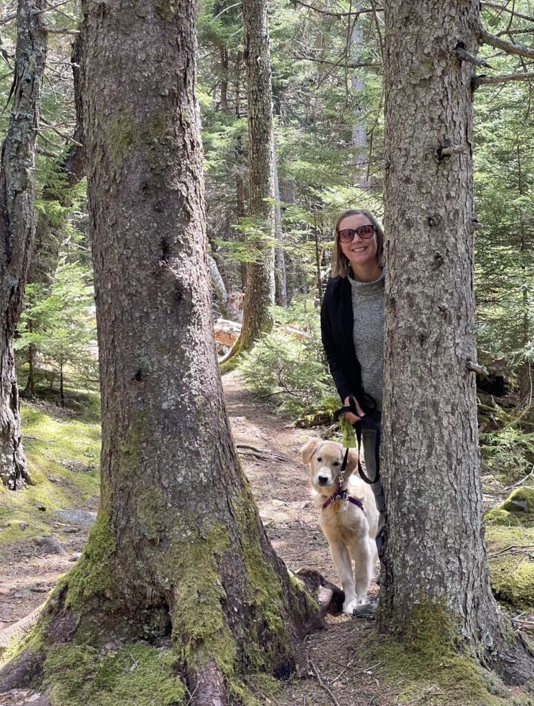 Picture of female with puppy peeking from behind a tree on a trail in Acadia National Park during Spring